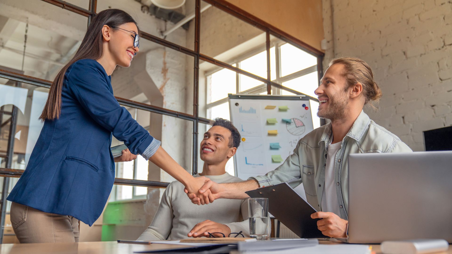 Usměvaví kolegové si podávají ruce v moderním kancelářském prostředí.